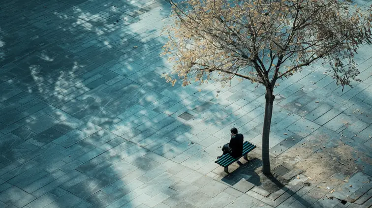 A high-angle shot captures a lone figure sitting on a bench in an empty urban park